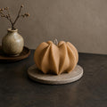 A sculptural pumpkin-shaped candle in a warm tan color sits on a round stone coaster on a dark tabletop. In the background, a small speckled ceramic vase with dried branches is visible against a textured neutral wall. Wavy Ribbed Candle, Soy Wax Pillar Candle, Swirl Candle by Votive Candle Co