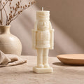 A white ceramic nutcracker candle stands on a tabletop in a styled interior setting with a beige vase, dried branches, and a wooden dish in the background. Hand-Poured Nutcracker Candle: Sculptural Soy Wax Christmas Decor