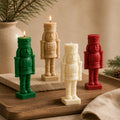 Four nutcracker-shaped candles in green, beige, cream, and red, arranged on a wooden tray with a warm neutral background and holiday greenery.Hand-Poured Nutcracker Candle: Sculptural Soy Wax Christmas Decor