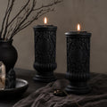 Gothic Pillar Candle, Dark Academia Decor, Victorian Pillar Candle. Two black ornate pillar candles with lit flames, styled on a dark tabletop with draped fabric, a ceramic vase with bare branches, and small decorative crystals.