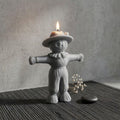 A gray scarecrow-shaped candle holder with a lit candle on top, set on a dark textured mat against a neutral gray background. Scarecrow Soy Wax Candle – Halloween Fall Decor