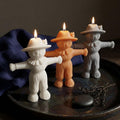 Three scarecrow-shaped candle holders shaped like people, lit with small flames, displayed on a dark tray with a blue cloth and small decorative branch in the background. Scarecrow Soy Wax Candle – Halloween Fall Decor