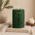 A dark green cylindrical holiday candle with a white wick, embossed pine branches and pinecones, and the word 'Joy' in gold script. The candle is styled on a stone block with a beige cloth, a ceramic vase with pine branches, and a small decorative ornament in a warm neutral setting. Christmas Soy Pillar Candle, Christmas Ornament, Christmas Candle Gift