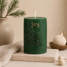 A dark green cylindrical holiday candle with a white wick, embossed pine branches and pinecones, and the word 'Joy' in gold script. The candle is styled on a stone block with a beige cloth, a ceramic vase with pine branches, and a small decorative ornament in a warm neutral setting. Christmas Soy Pillar Candle, Christmas Ornament, Christmas Candle Gift