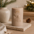 A beige holiday candle with embossed pine branches and the word 'Joy' in gold lettering, displayed on a stone coaster with a pinecone and warm blurred background lights. Christmas Soy Pillar Candle, Christmas Ornament, Christmas Candle Gift