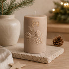 A beige holiday candle with embossed pine branches and the word 'Joy' in gold lettering, displayed on a stone coaster with a pinecone and warm blurred background lights. Christmas Soy Pillar Candle, Christmas Ornament, Christmas Candle Gift