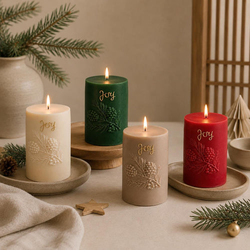 Four embossed holiday candles with the word 'Joy' in gold script, displayed in cream, green, taupe, and red on a styled tabletop with pine branches and neutral decor.