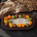 Fall Dough Bowl Candle, Handmade Soy Wax Wooden Bowl Candle. An oval dark brown candle holder with a white wax center and colorful fall-themed decorative elements around the edge, including orange pumpkins, green gourds, yellow leaves, acorns, and brown acorns, styled on a dark surface with a beige linen cloth and dried flowers in the background.