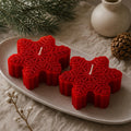 Two red snowflake-shaped candles with white wicks, displayed on a speckled beige ceramic tray with a pinecone, a small vase, and evergreen branches in the background. Snowflake Candle Gift Set, Handmade Soy Wax Christmas Candles, Winter Decor