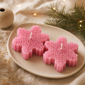 Two pink snowflake-shaped candles arranged on a beige ceramic plate, styled with a small vase, dried flowers, and evergreen branches with warm string lights in the background. Snowflake Candle Gift Set, Handmade Soy Wax Christmas Candles, Winter Decor