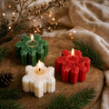 Three snowflake-shaped candle holders in green, red, and white, each holding a lit tealight candle, arranged on a rustic wooden board with evergreen branches, a pinecone, and soft beige fabric in the background.