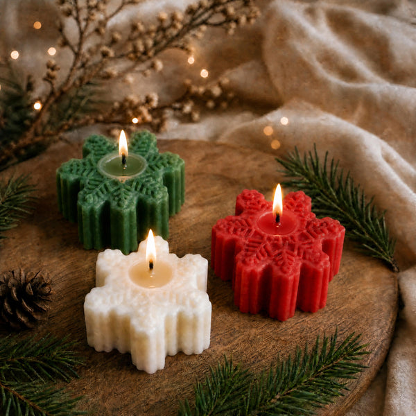 Three snowflake-shaped candle holders in green, red, and white, each holding a lit tealight candle, arranged on a rustic wooden board with evergreen branches, a pinecone, and soft beige fabric in the background.