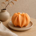 An orange sculptural candle with a ribbed, pumpkin-like surface sits lit on a small beige ceramic plate on a wooden table. A matching vase with dried branches and a beige cloth are softly blurred in the background. Wavy Ribbed Candle, Soy Wax Pillar Candle, Swirl Candle by Votive Candle Co