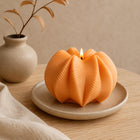 An orange sculptural candle with a ribbed, pumpkin-like surface sits lit on a small beige ceramic plate on a wooden table. A matching vase with dried branches and a beige cloth are softly blurred in the background. Wavy Ribbed Candle, Soy Wax Pillar Candle, Swirl Candle by Votive Candle Co
