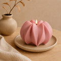 A pink sculptural candle with a flower-like ribbed shape sits lit on a small beige ceramic plate, placed on a wooden table with a beige vase and cloth in the background. Wavy Ribbed Candle, Soy Wax Pillar Candle, Swirl Candle by Votive Candle Co