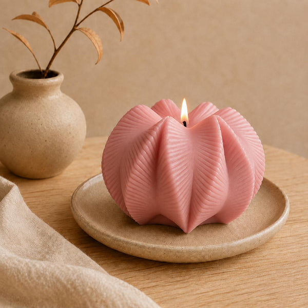 A pink sculptural candle with a flower-like ribbed shape sits lit on a small beige ceramic plate, placed on a wooden table with a beige vase and cloth in the background. Wavy Ribbed Candle, Soy Wax Pillar Candle, Swirl Candle by Votive Candle Co