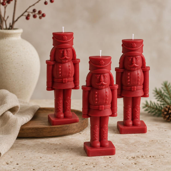 Three red nutcracker-shaped candles standing on a beige stone surface, with a cream vase, draped fabric, and small evergreen sprigs in the background. Hand-Poured Nutcracker Candle: Sculptural Soy Wax Christmas Decor