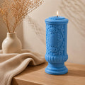 Gothic Pillar Candle, Dark Academia Decor, Victorian Pillar Candle. A blue ornate pillar candle with a single lit wick sits on a wooden table next to a beige textured throw blanket and a small ceramic vase with dried stems.