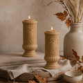 Gothic Pillar Candle, Dark Academia Decor, Victorian Pillar Candle. Two ornate beige pillar candles with carved details and lit flames, styled on a rustic wooden surface with a linen cloth, dried leaves, a ceramic bowl, and a vase in the background.