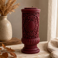 Gothic Pillar Candle, Dark Academia Decor, Victorian Pillar Candle. A dark red ornate pillar candle with intricate scrollwork relief patterns, displayed on a stone surface next to a beige ceramic vase with dried grasses.