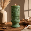 Gothic Pillar Candle, Dark Academia Decor, Victorian Pillar Candle. A green ornate pillar candle with a lit flame, styled on a wooden table with dried leaves, a ceramic vase, and a bowl in a warm, rustic setting.