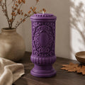 Gothic Pillar Candle, Dark Academia Decor, Victorian Pillar Candle. A purple ornate pillar candle with a detailed relief pattern and a small white wick, displayed on a dark wooden table with a beige ceramic vase, dried branches, and a brown cloth in the background.
