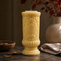 Gothic Pillar Candle, Dark Academia Decor, Victorian Pillar Candle. A tall, ornate yellow pillar candle with intricate carved scrollwork and a central oval medallion, displayed on a rustic wooden surface with a ceramic vase, cloth, and dried leaves in the background.