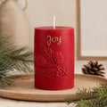 A red cylindrical holiday candle with an embossed pine branch and berry design, featuring the word 'Joy' in gold script, displayed on a wooden tray with pine branches and a pinecone. Christmas Soy Pillar Candle, Christmas Ornament, Christmas Candle Gift