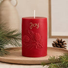 A red cylindrical holiday candle with an embossed pine branch and berry design, featuring the word 'Joy' in gold script, displayed on a wooden tray with pine branches and a pinecone. Christmas Soy Pillar Candle, Christmas Ornament, Christmas Candle Gift