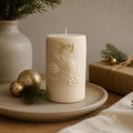 A cream-colored holiday candle with embossed pinecones and the word 'Joy' in gold, styled on a ceramic tray with gold ornaments, pine branches, and a wrapped gift box in a warm indoor setting. Christmas Soy Pillar Candle, Christmas Ornament, Christmas Candle Gift