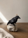 A black raven candle perched on a white skull-shaped base, displayed on a wooden surface against a neutral beige wall. Raven Skull Candle – Large Handmade Gothic Sculptural Candle, Dark Academia Halloween Decor