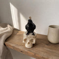 A black bird candle perched on top of an off-white skull candle holder, styled on a rustic wooden shelf next to a beige ceramic cup and draped fabric. Raven Skull Candle – Large Handmade Gothic Sculptural Candle, Dark Academia Halloween Decor