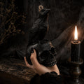 A black raven candle perched on a black skull, held in a hand with black nail polish against a dark, moody background with a lit black candle. Raven Skull Candle – Large Handmade Gothic Sculptural Candle, Dark Academia Halloween Decor