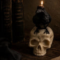 A gothic-themed candle shaped like a black raven perched on a skull, lit with a small flame, set on a dark wooden surface next to old books. Raven Skull Candle – Large Handmade Gothic Sculptural Candle, Dark Academia Halloween Decor