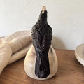A black bird figurine with textured feathers perched on a round, matte off-white base, set on a wooden surface against a light wall with draped fabric in the background. Raven Skull Candle – Large Handmade Gothic Sculptural Candle, Dark Academia Halloween Decor