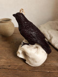 A dark brown raven figurine perched on a white skull, with a small beige hanging loop at the top, displayed on a rustic wooden surface with a neutral background. Raven Skull Candle – Large Handmade Gothic Sculptural Candle, Dark Academia Halloween Decor