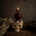A dark brown crow-shaped candle with a lit flame sits on top of an off-white skull, placed on a rustic wooden surface with a draped cloth and an old book in a dim, moody setting. Raven Skull Candle – Large Handmade Gothic Sculptural Candle, Dark Academia Halloween Decor