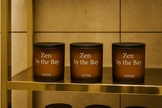 Three 'Zen by the Bay' candles on a gold shelf against a beige tiled wall.