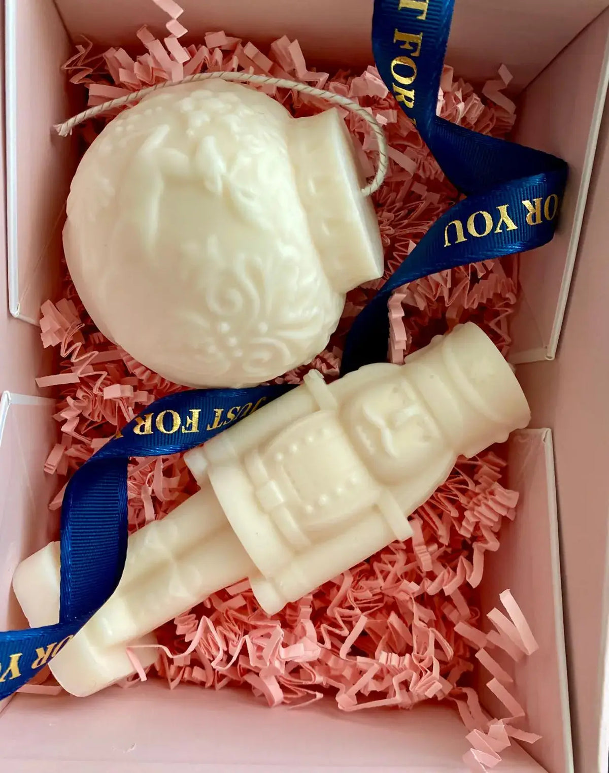 Two white nutcracker and ornament scented candles nestled in pink shredded paper, elegantly packaged with a ribbon.