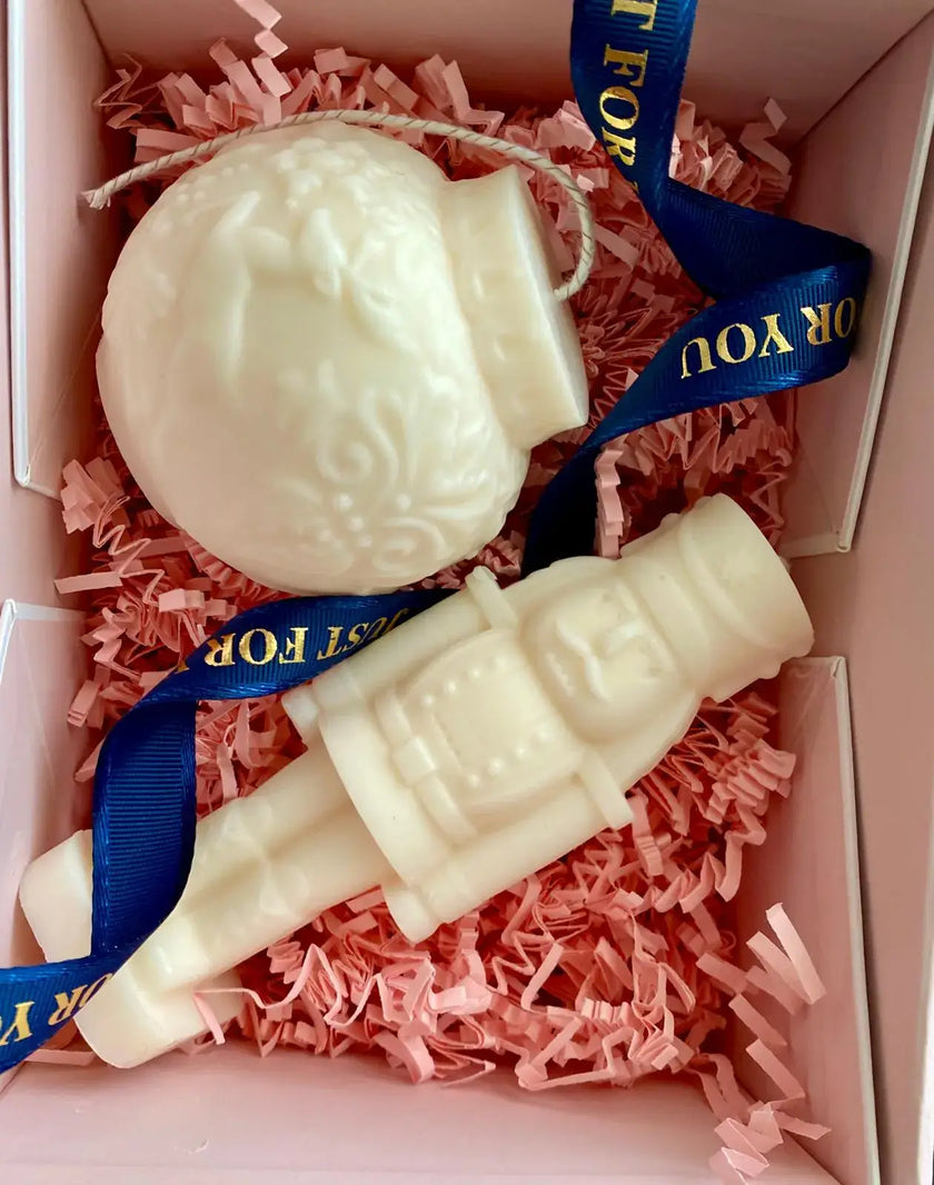 Two white nutcracker and ornament scented candles nestled in pink shredded paper, elegantly packaged with a ribbon.