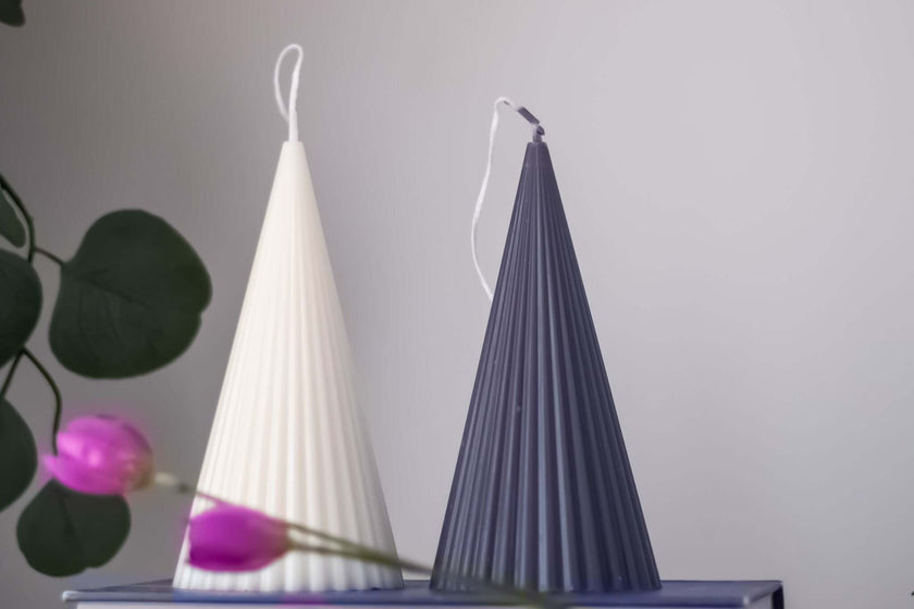 Two cone-shaped handmade soy wax candles, one white and one dark gray, on a table with flowers.