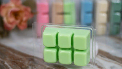 Pack of green wax melts in a clear plastic packaging on a wooden surface with blurred colorful candles in the background.