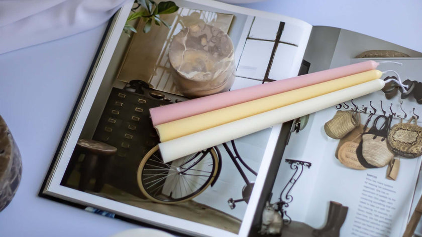 Three handmade taper candles in pastel colors placed on an open magazine, showcasing their elegant design.